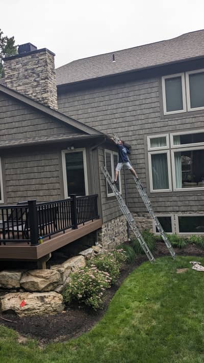 House exterior with gray shingle siding, ladder against wall, and stone chimney.