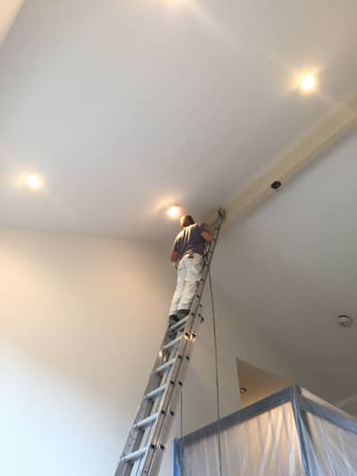 White painted ceiling with recessed lighting fixtures and painter on ladder mid-installation.