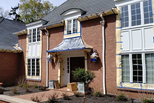 A brick house with its entry canopy tented in plastic and window trim masked in paper and blue tape.
