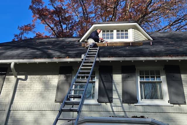 A painter on an extension ladder working on a dormer above a painted brick house, autumn trees behind.